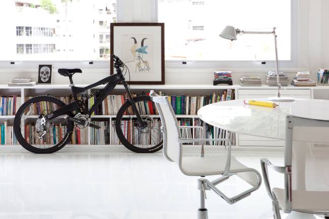 A imagem mostra uma bicicleta apoiada no aparador da sala. No primeiro plano uma mesa com uma cadeira.