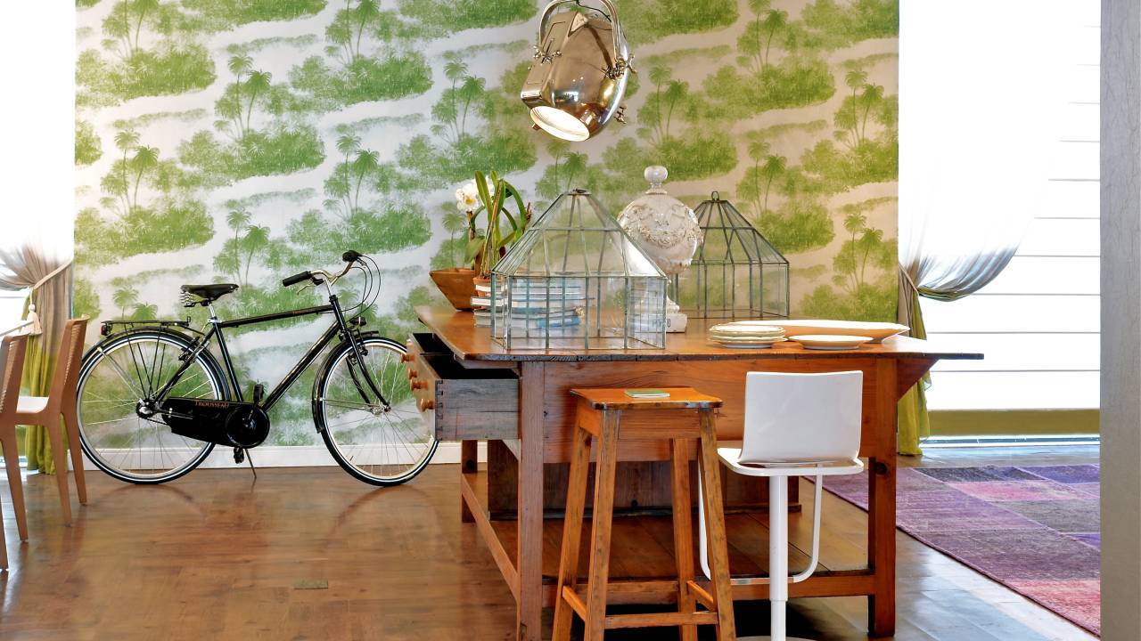 A imagem mostra no uma bicicleta apoiada no chão da sala de estar. No primeiro plano uma mesa de madeira com objetos de decoração.