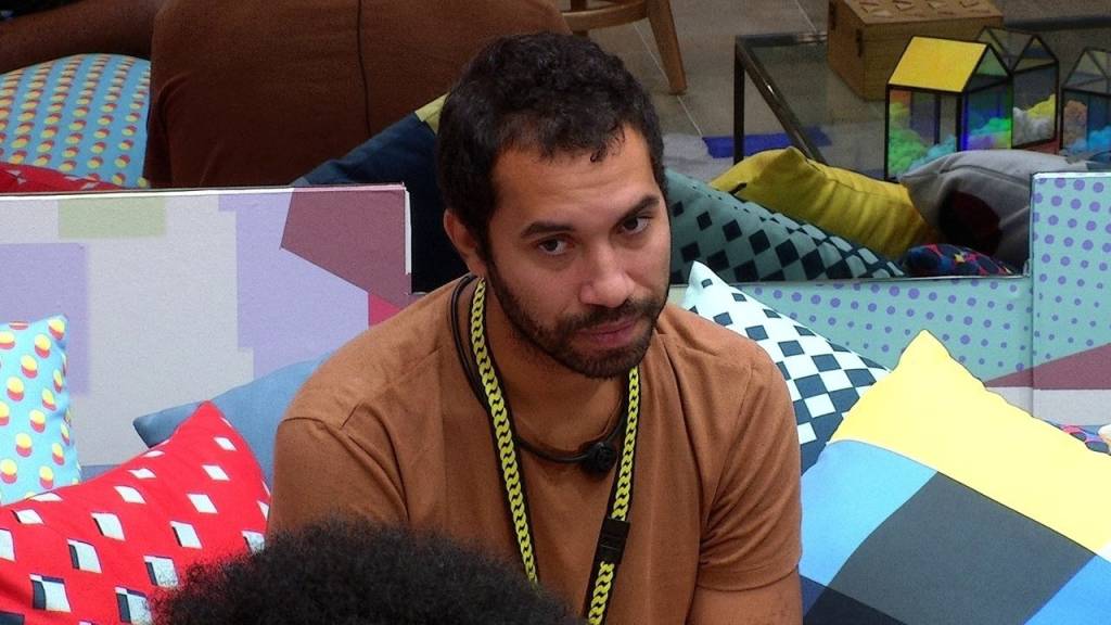 A imagem mostra o participante GIlberto, do Big Brother Brasil, de camiseta marrom, sentado numa cama