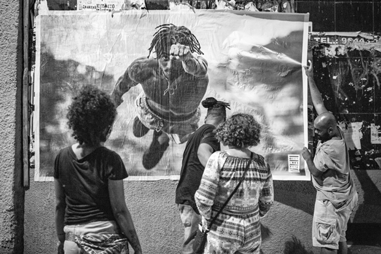 Pessoas em frente a um lambe-lambe na rua. A gravura mostra um homem negro voando no estilo Super Homem