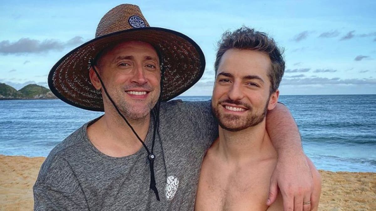 A imagem mostra o ator Paulo Gustavo, de chapeu, abraçaado ao marido, Thales Bretas, sem camisa. Os dois estão numa praia