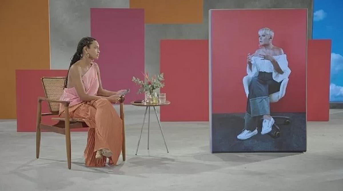 A imagem mostra Taís Araujo e Xuxa, a atriz sentada numa cadeira no cenário do GNT, Xuxa pelo telão