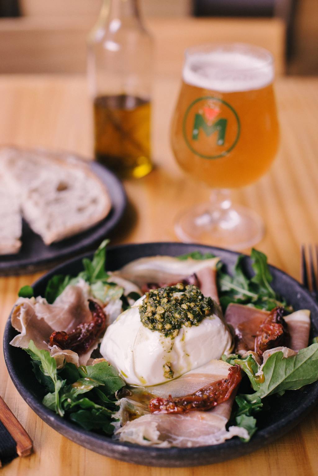 A imagem mostra uma salada de presunto cru, burrata e folhas, copo de chope e pão de fermentação natural