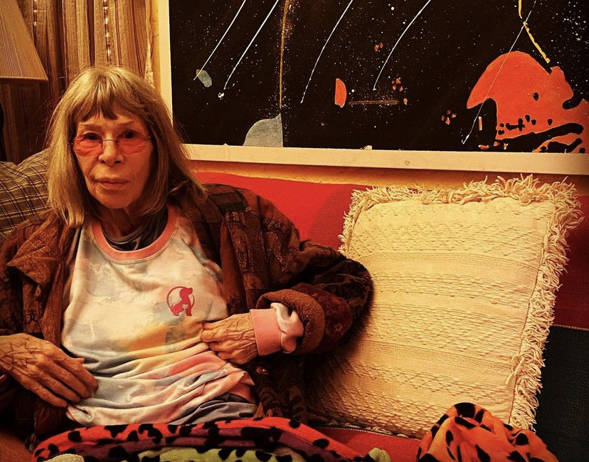 A imagem mostra Rita Lee sentada num sofá vermelho