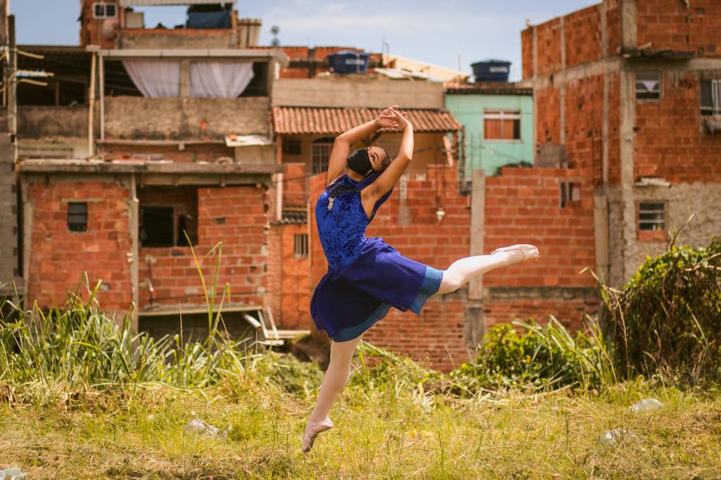 Bailarina, de máscara de segurança, faz movimento de dança em meio a uma favela