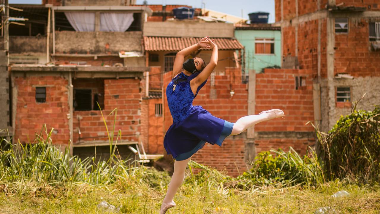 Bailarina, de máscara de segurança, faz movimento de dança em meio a uma favela