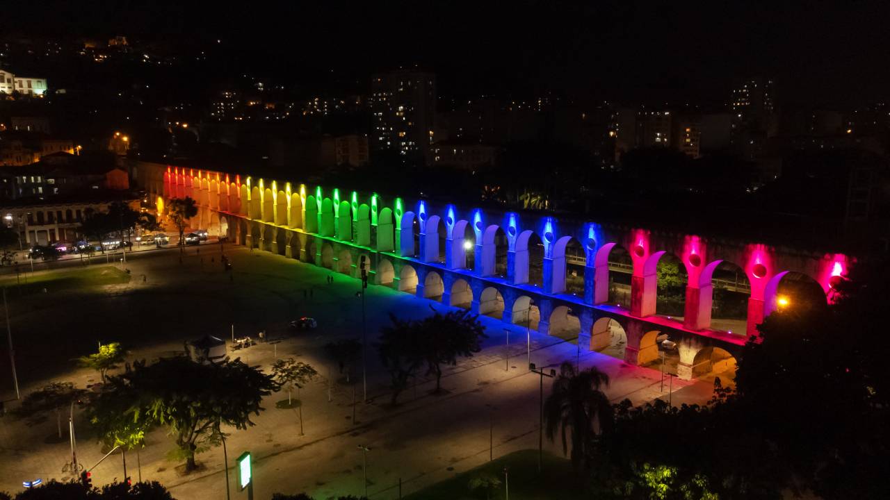 Imagem mostra os Arcos da Lapa à noite iluminados com as cores do arco-íris