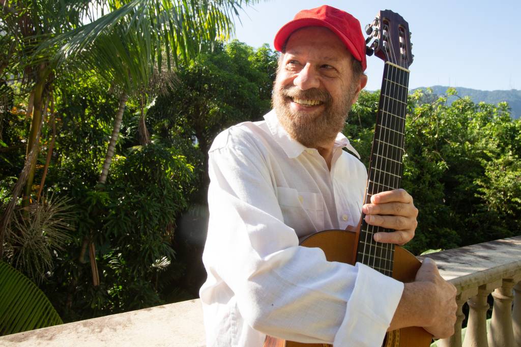 João Bosco de boné, segurando um violão, numa varanda arborizada