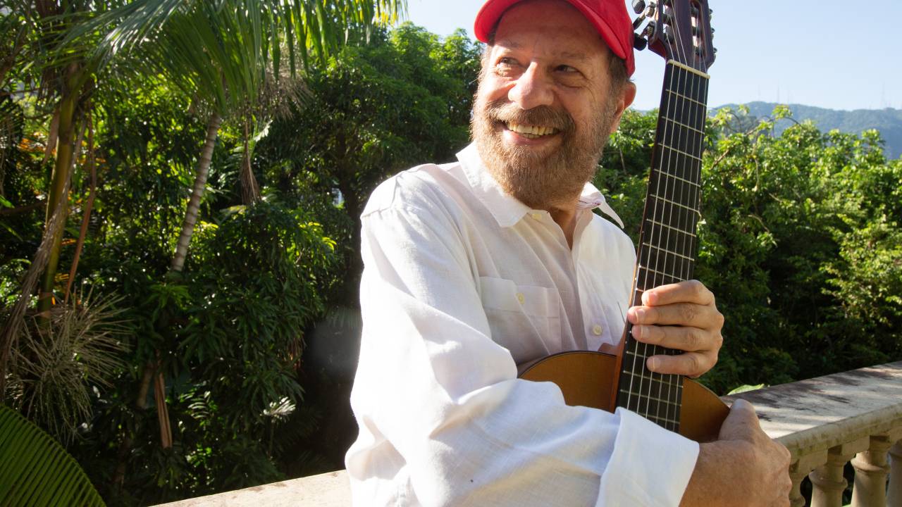 João Bosco de boné, segurando um violão, numa varanda arborizada