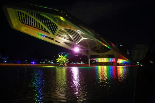 Imagem mostra Museu do Amanhã à noite iluminado com as cores da bandeira LGBTI+