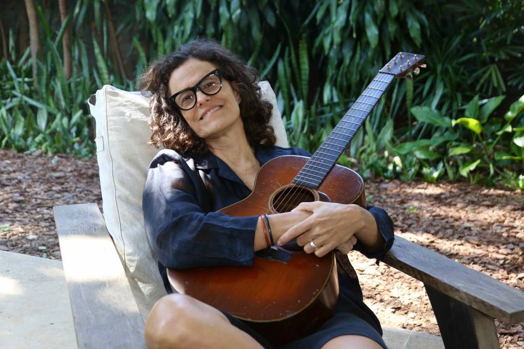 Zélia Duncan sentada numa poltrona, num jardim, segurando um violão, sorrindo de óculos