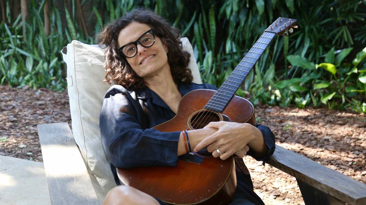 Zélia Duncan sentada numa poltrona, num jardim, segurando um violão, sorrindo de óculos