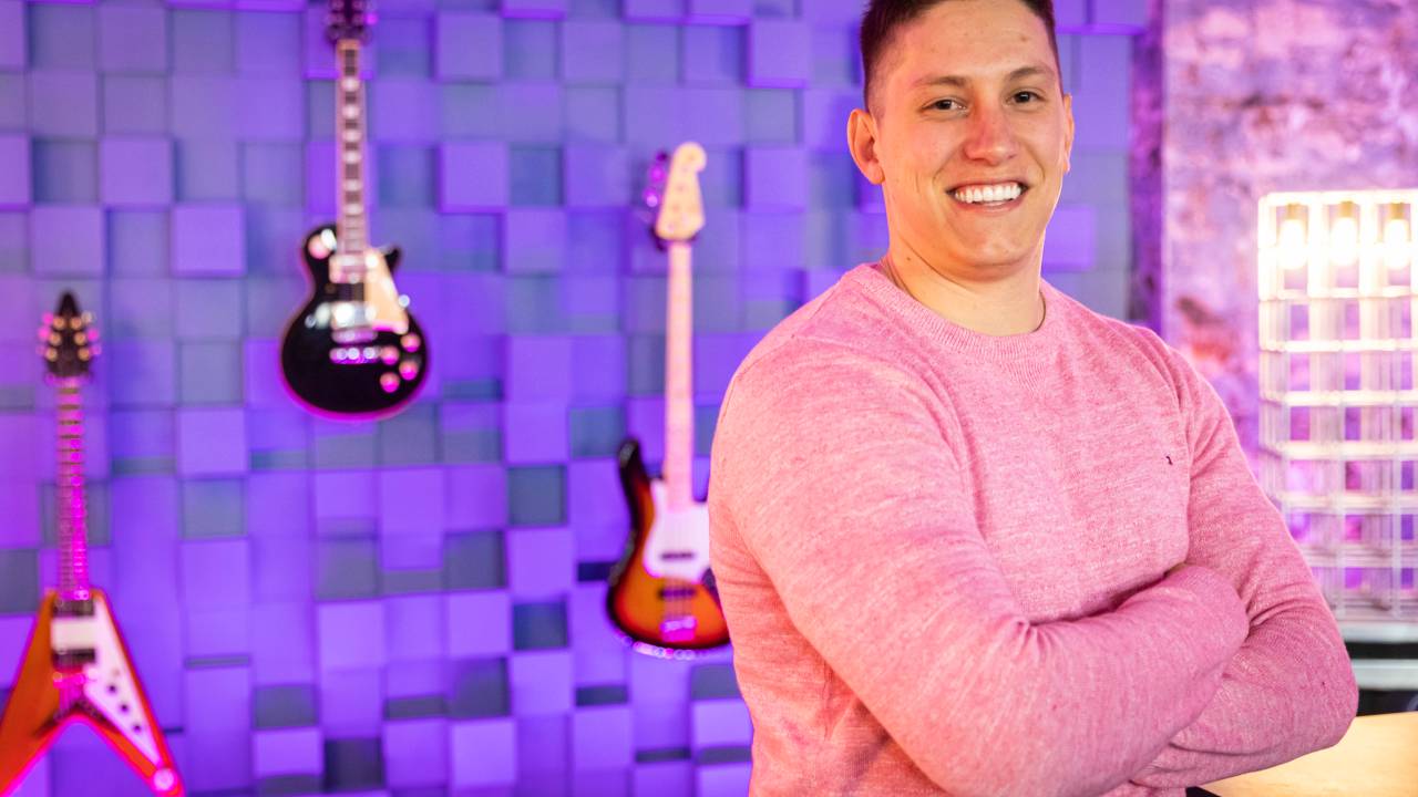 Jakson Follmann de camisa rosa, braços cruzados e guitarras ao fundo