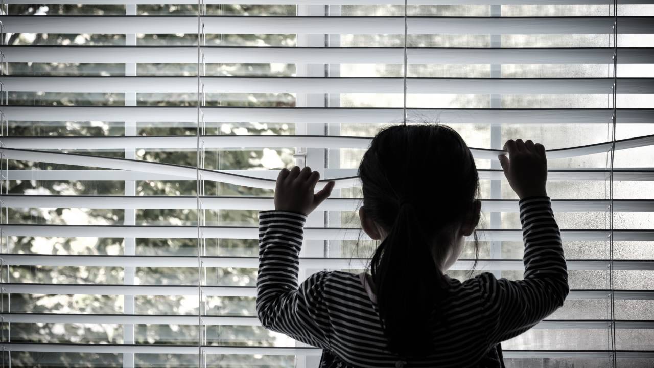 Menina levanta a persiana de uma janela para ver a vista do lado de fora.
