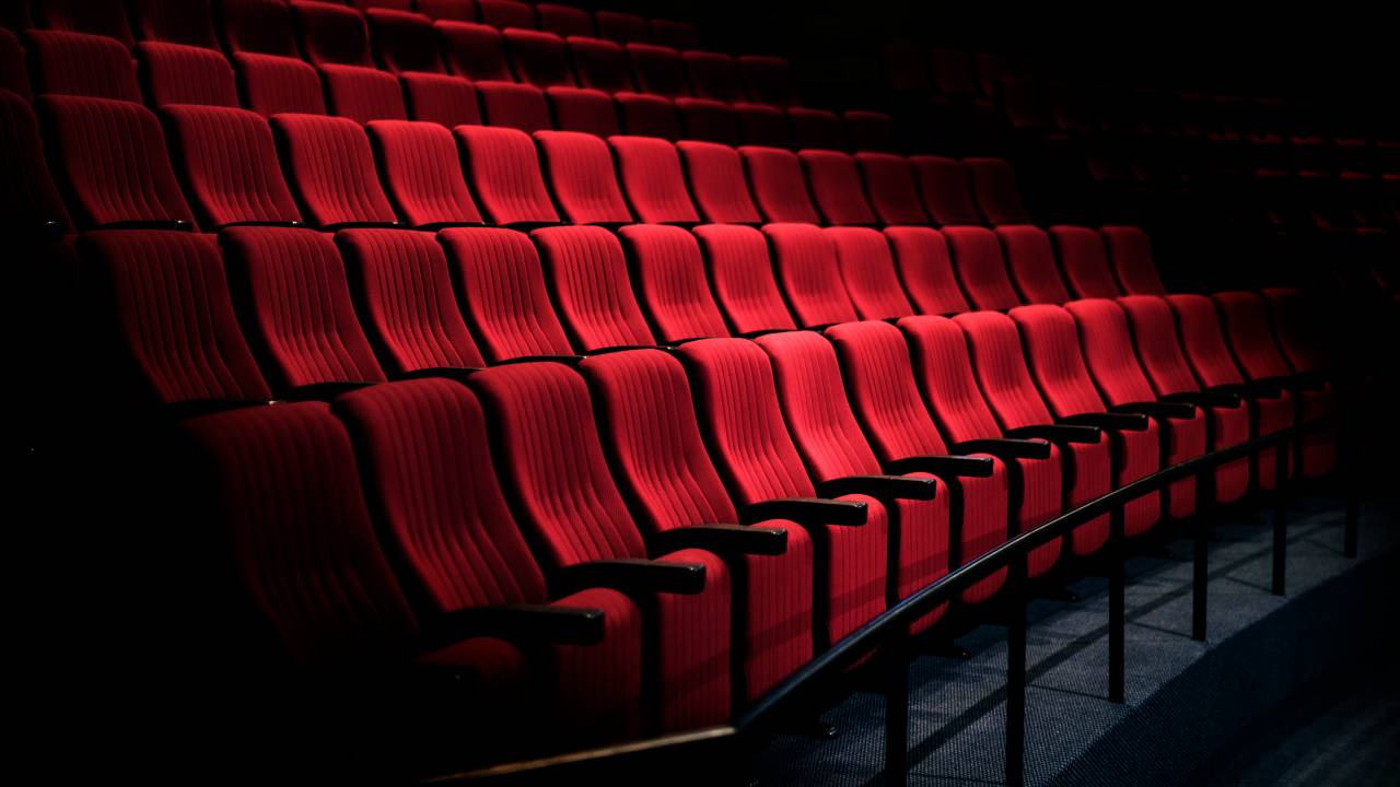 Cadeiras de teatro vermelhas estão vazias e iluminadas por um foco de luz do teatro