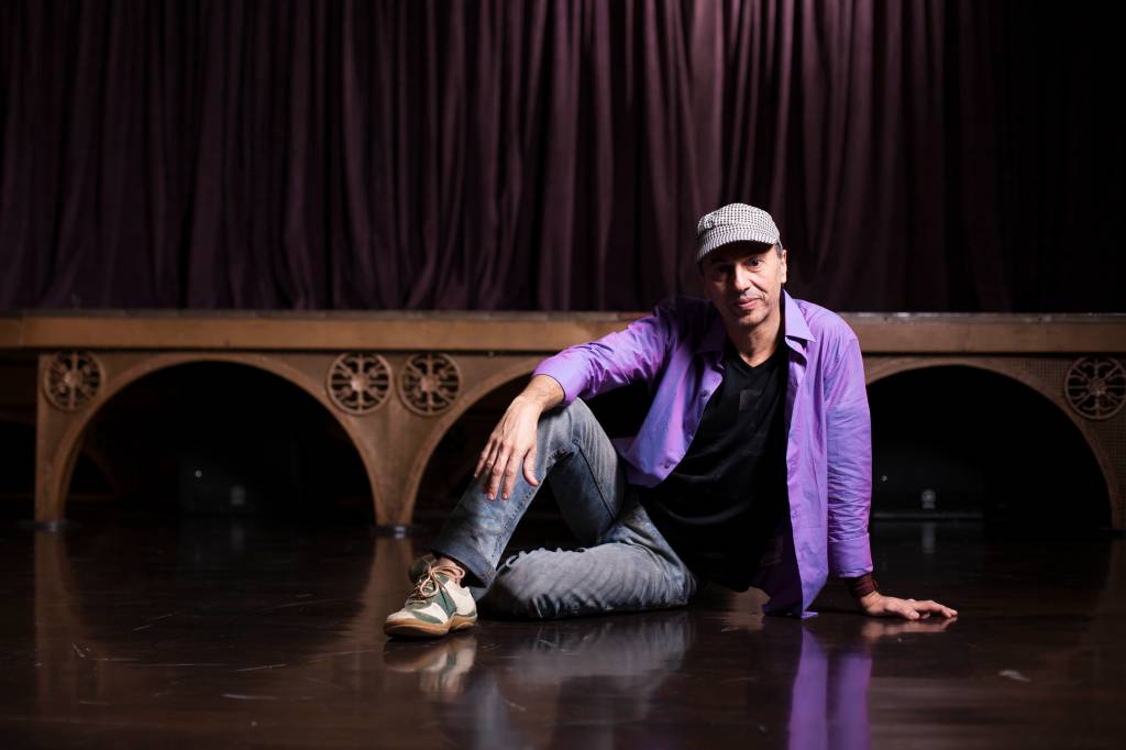 Zeca Baleiro sentado no chão de um palco, de boné e camisa lilás