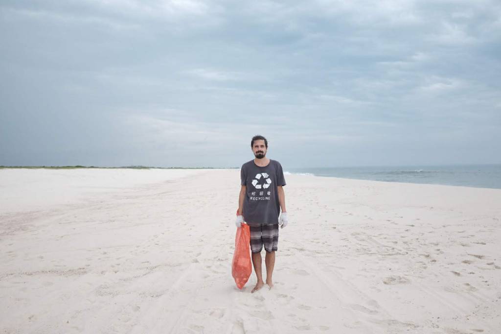 Foto mostra homem adulto segurando sacola plástica vermelha em uma praia
