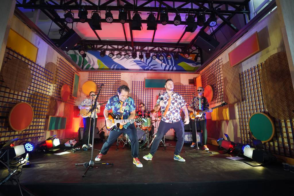 Foto mostra mostra banda tocando em um palco todo colorido