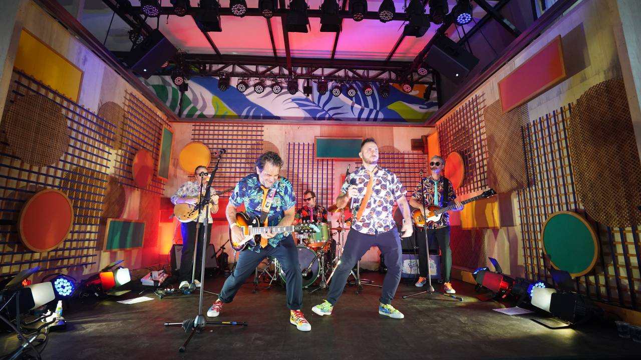 Foto mostra mostra banda tocando em um palco todo colorido