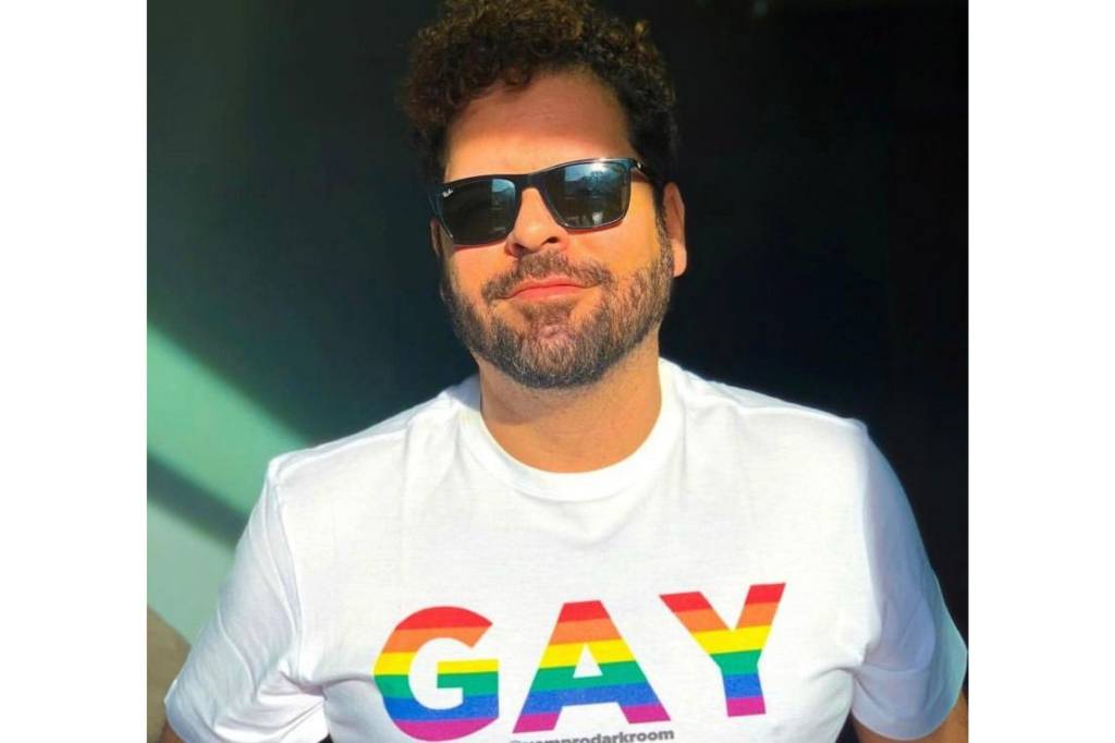 Imagem mostra homem com barba usando óculos escuros e uma camisa branca com a palavra gay nas cores do arco-íris.