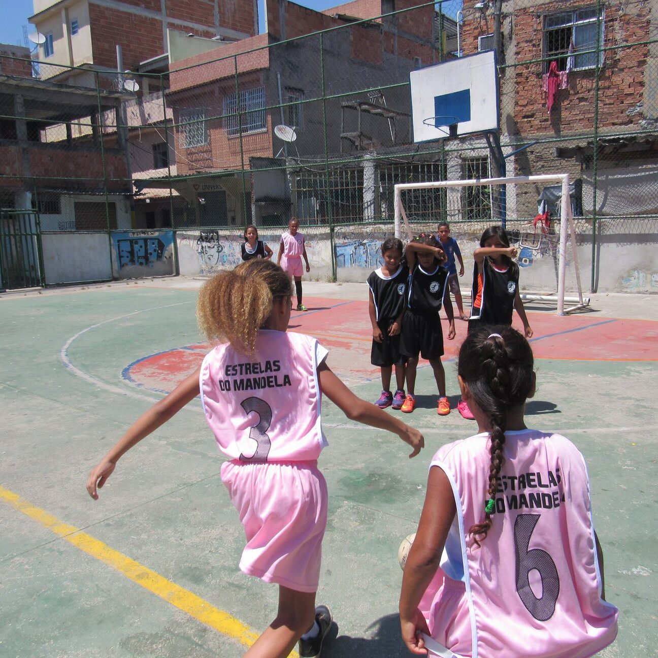 Imagem mostra meninas com uniforme rosa claro jogando futebol em uma quadra da comunidade
