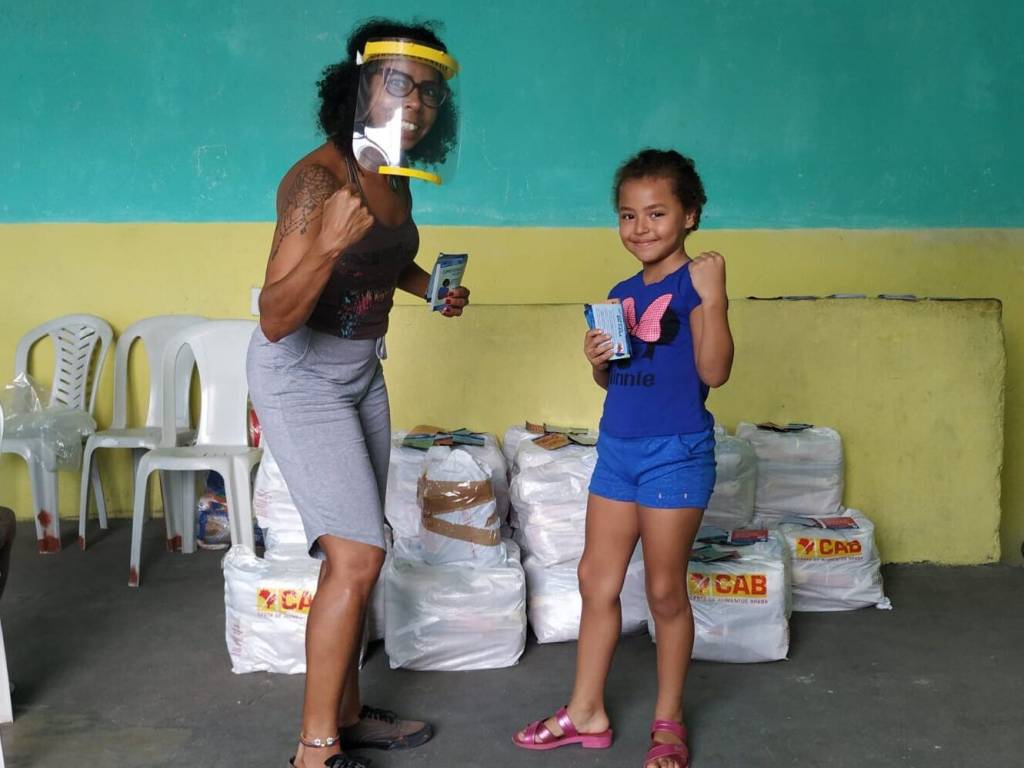 Imagem mostra uma mulher e uma menina em frente a cestas básicas