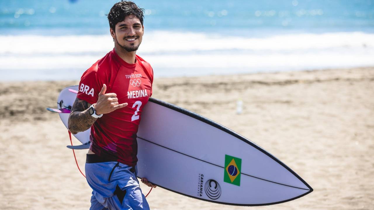 A imagem mostra Gabriel Medina com a prancha de surfe fazendo sinal de positivo com a mão