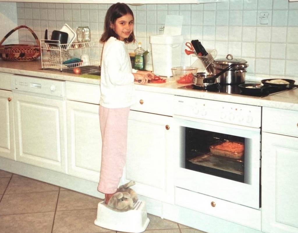 Foto mostra menina cozinhando em cima de um banquinho branco