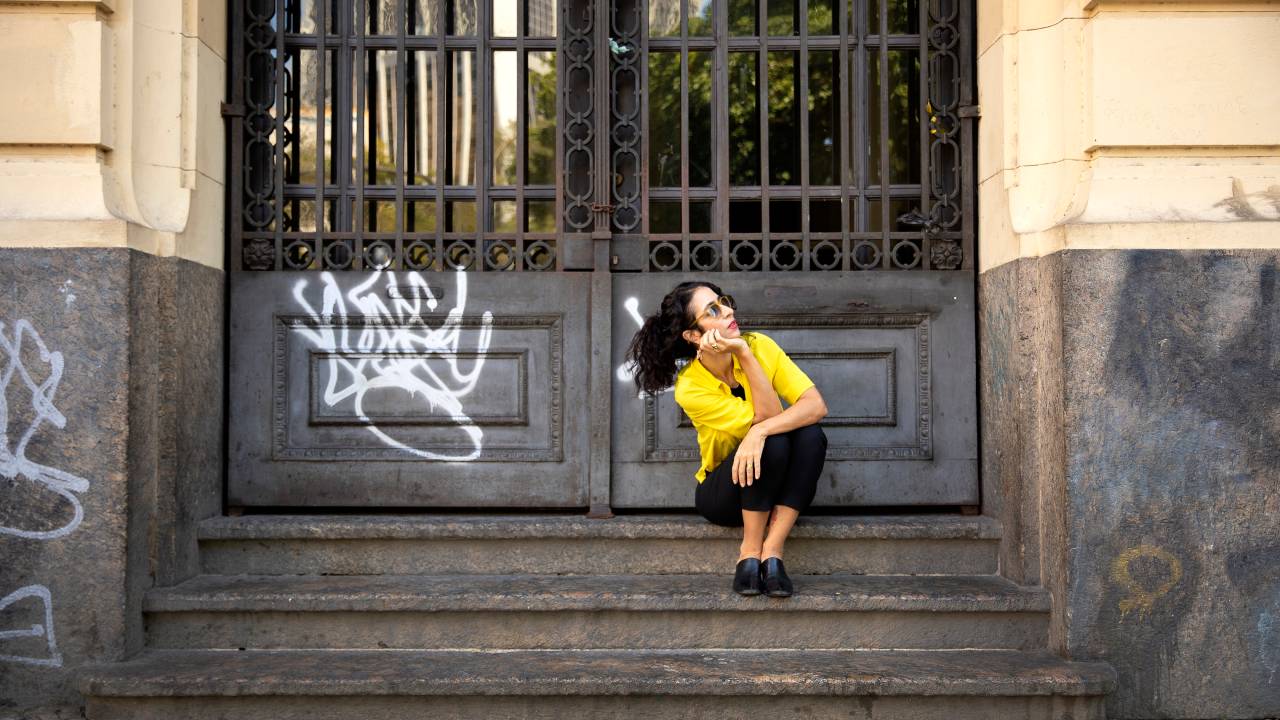 Marisa Monte sentada numa escadinha, na frente de um portão pichado, virada para o lado