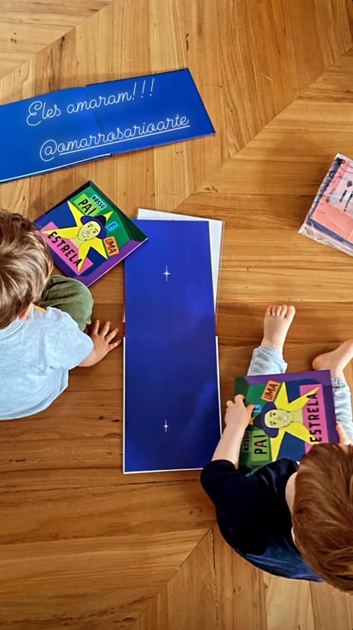 Imagem mostra duas crianças penas segurando dois livros infantis, sentadas em um chão de madeira