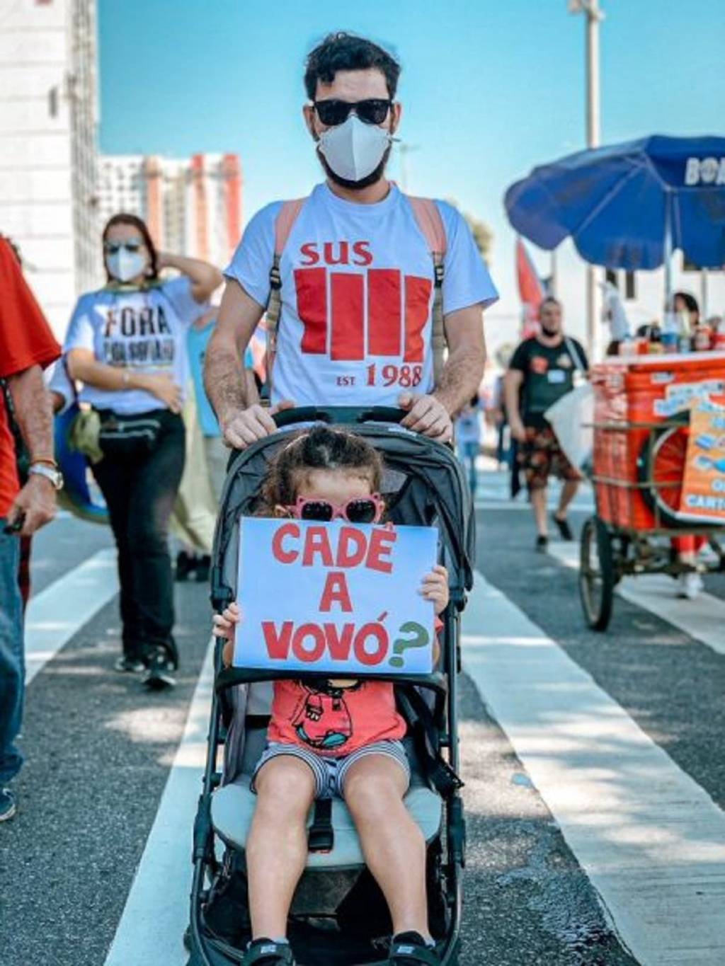 A imagem mostra uma menina pequena no carrinho de bebê segurando um cartaz onde se lê 