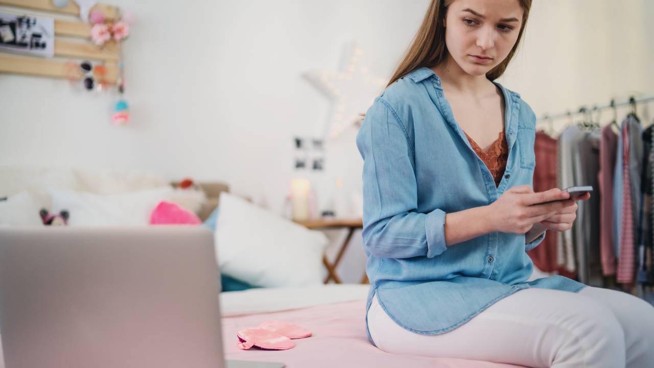 Jovem em seu quarto cercada por celular e computador.