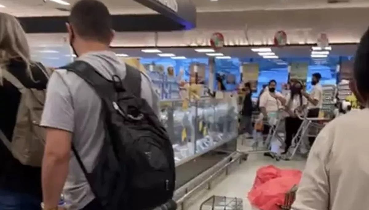 A imagem mostra um corpos coberto com um pano vermelho no corredor de um supermercado, com algumas pessoas em volta