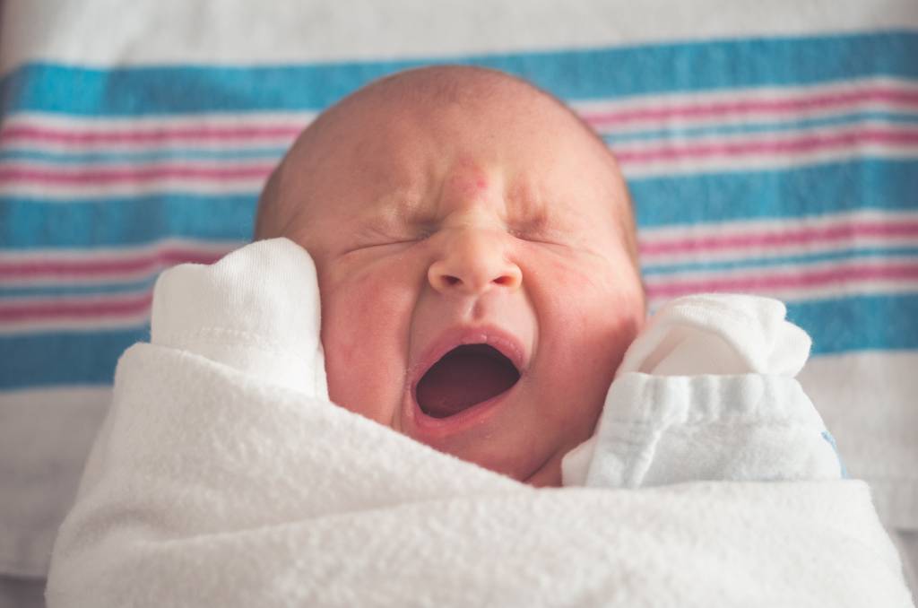 Bebê recém nascido com a boca aberta e todo enrolado com manta branca