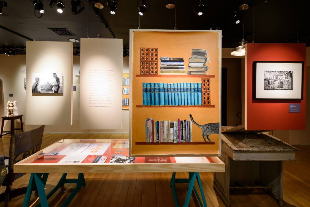 Exposição sobre Nise da Silveira no CCB, traz uma pintura de uma estante e uma mesa ao centro com alguns livros