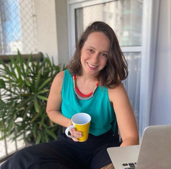 Foto mostra mulher branca com cabelo castanho na altura dos ombros sorrindo e segurando uma caneca. Ela está sentada em frente a um notebook branco. Ao fundo, há uma janela e um vaso de plantas.