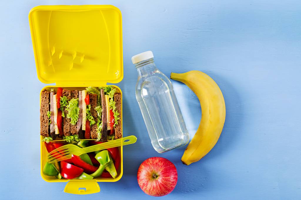 Foto mostra lancheira amarela com sanduíches e ao lado uma garrafa d'água, uma banana e uma maçã