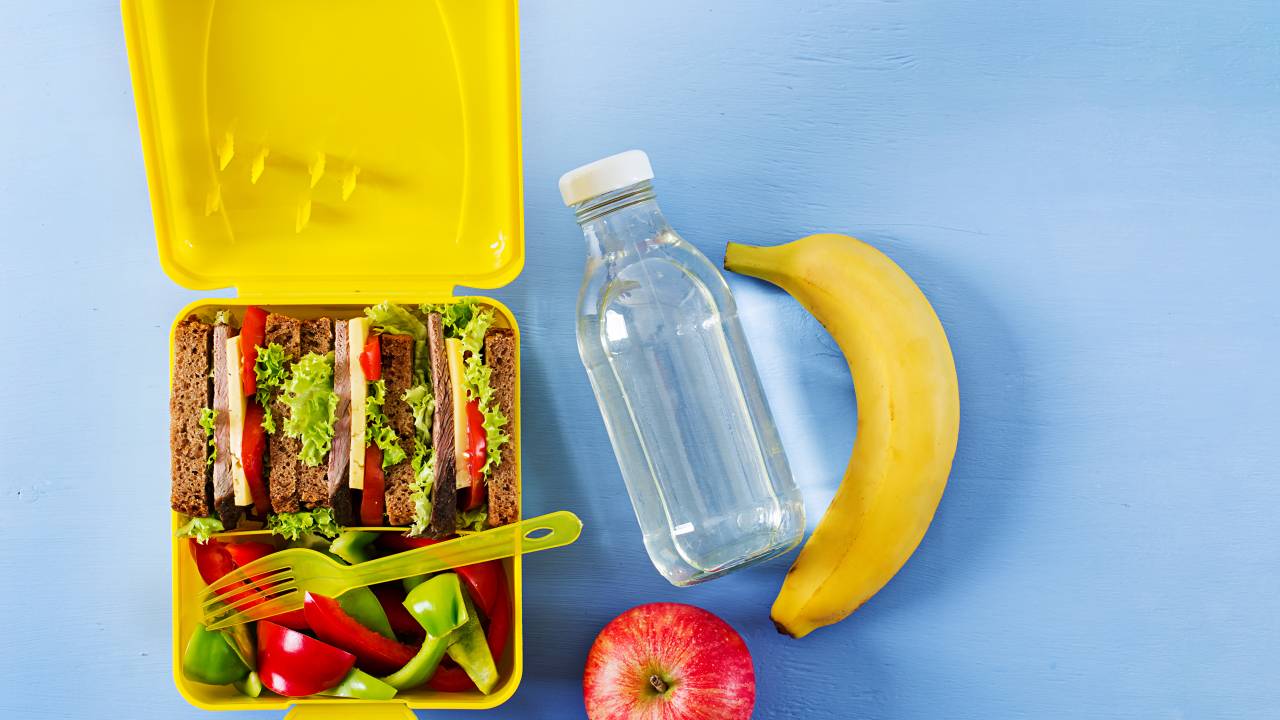Foto mostra lancheira amarela com sanduíches e ao lado uma garrafa d'água, uma banana e uma maçã
