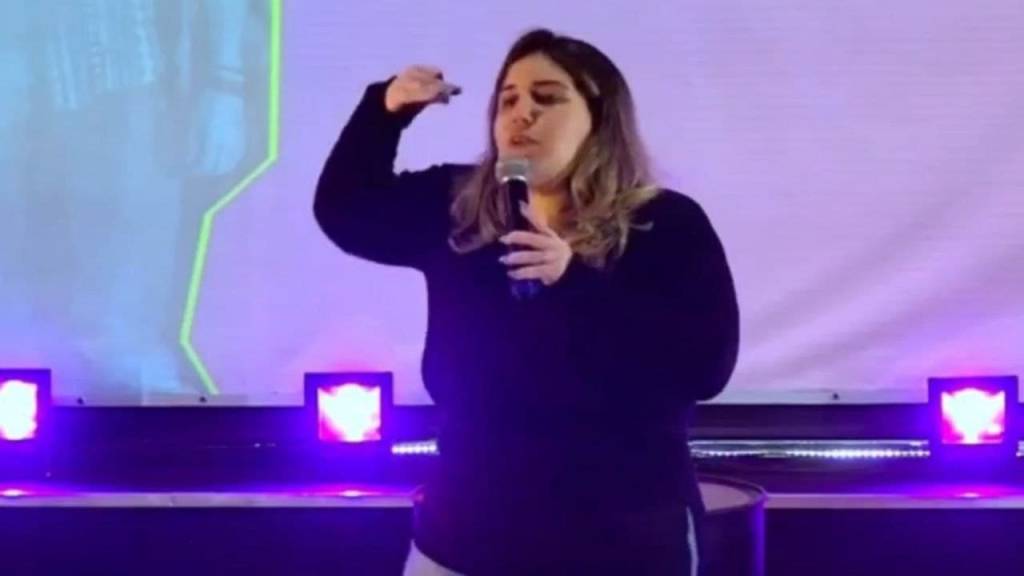 A imagem mostra uma mulher de casaco preto com o microfone na mão, gesticulando