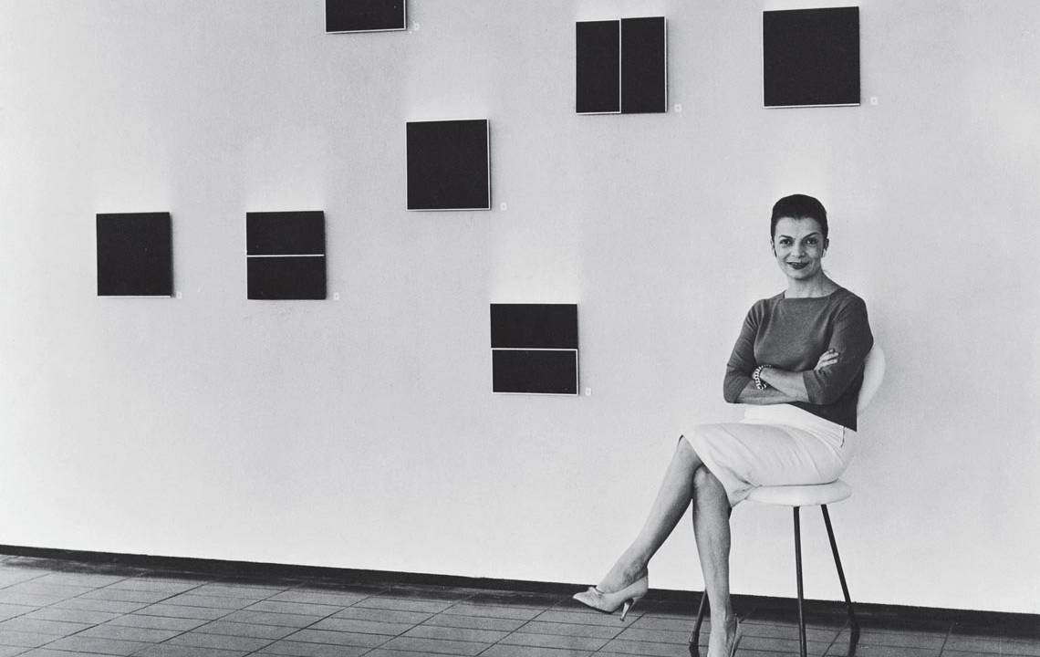 Artista plástica Lygia Clark sentada numa cadeira em frente a pinturas feitas por ela