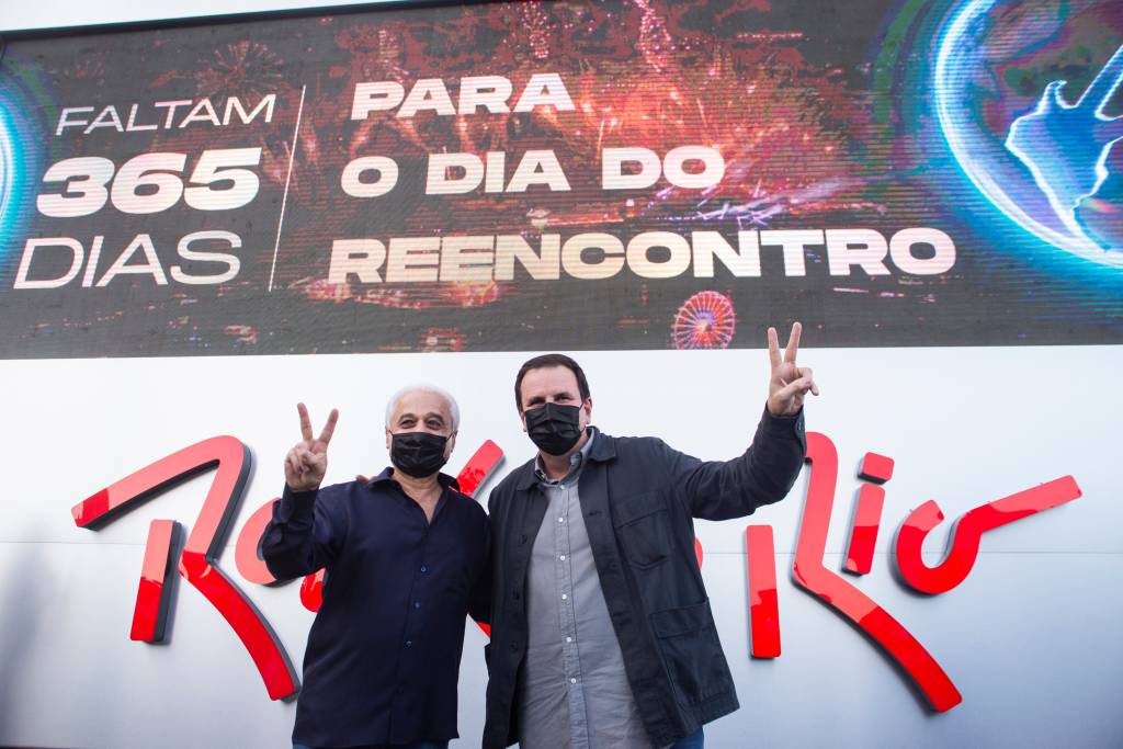 Roberto Medina e Eduardo Paes posam em frente ao relógio que marca a contagem regressiva para o Rock in Rio de 2022. Ele estão de mascara e fazem V com as mãos