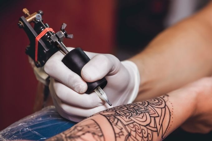 Uma mão com luva descartável segura uma maquina de tatuagem e faz um desenho em um braço branco.
