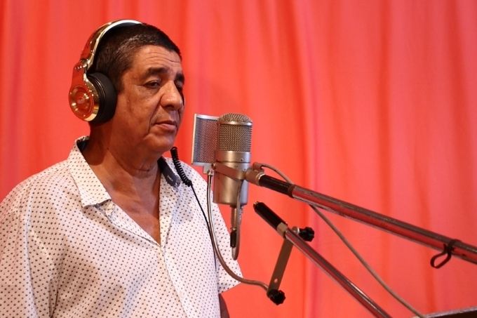Zeca Pagodinho veste uma blusa branca e usa fones no ouvido ao lado de um microfone de estúdio