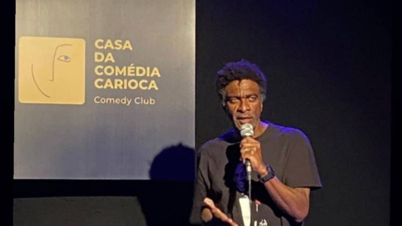 Humorista Helio de La Peña se apresenta com um microfone na mão, de camisa preta, em frente a uma placa que diz Clube da Comédia Carioca
