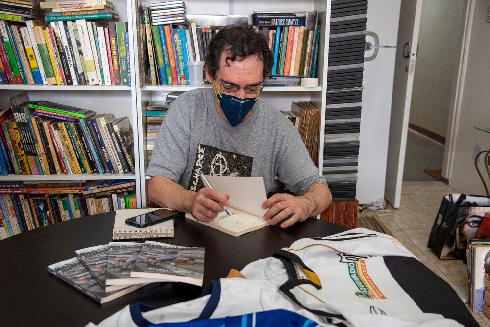 Kleber usa máscara facial e camisa cinza. Ele autografa um livro. Atrás dele há uma estante cheia de livros.