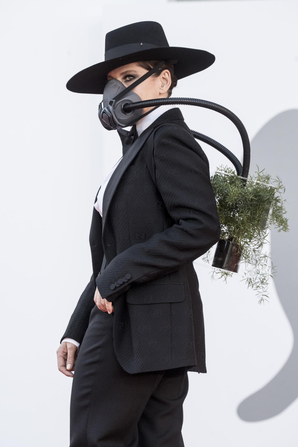 A imagem mostra a atriz Barbara Paz com uma máscara no rosto acoplada a um vaso de planta carregado nas costas