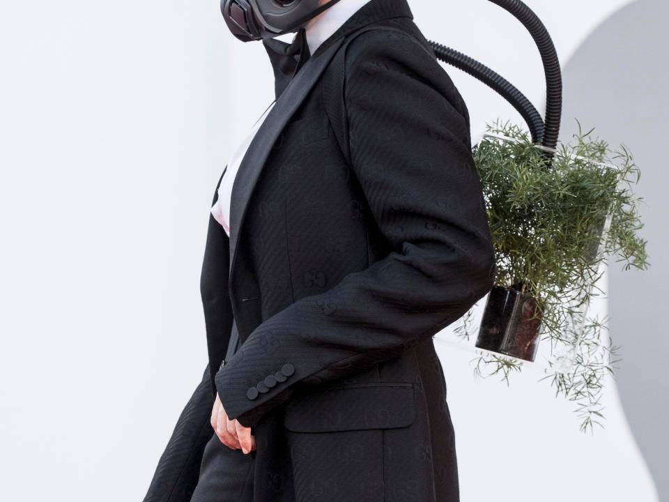 A imagem mostra a atriz Barbara Paz com uma máscara no rosto acoplada a um vaso de planta carregado nas costas
