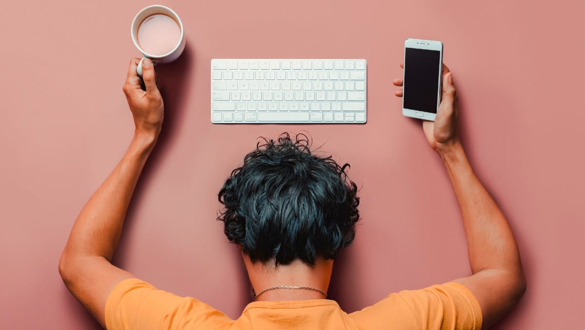 Homem com a cabeça sobre a mesa, diante de um teclado, com celular na mão e xícara de café.