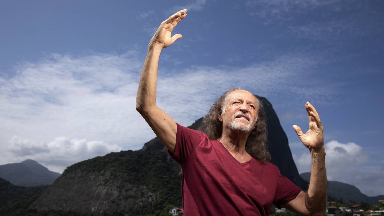 O cantor Alceu Valença veste blusa vinho e está com os braços abertos. Ao fundo, o Morro da Urca.