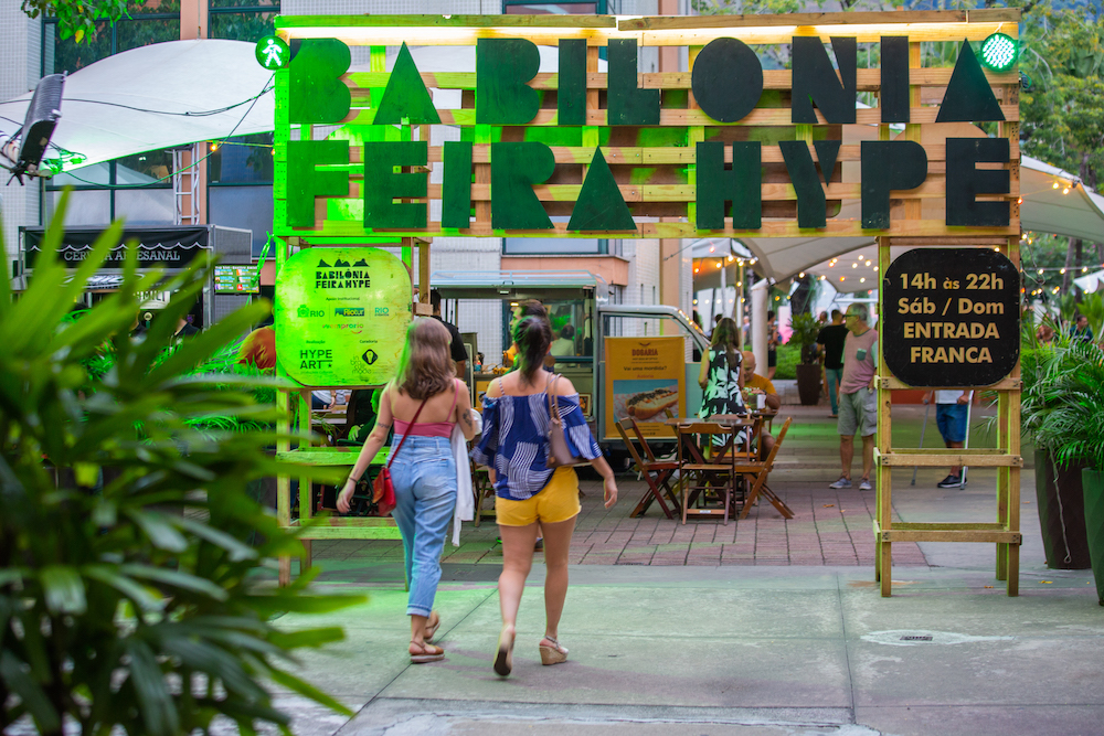 Uma placa escrito Babilônia Feira hype é iluminada por uma luz verde na entrada da feira e duas pessoas de costas estão entrando